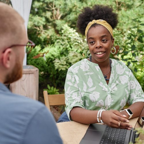 Medium shot of positive African American female landscape designer talking to male client discussing public park project, while sitting at table with laptop in sunlit garden